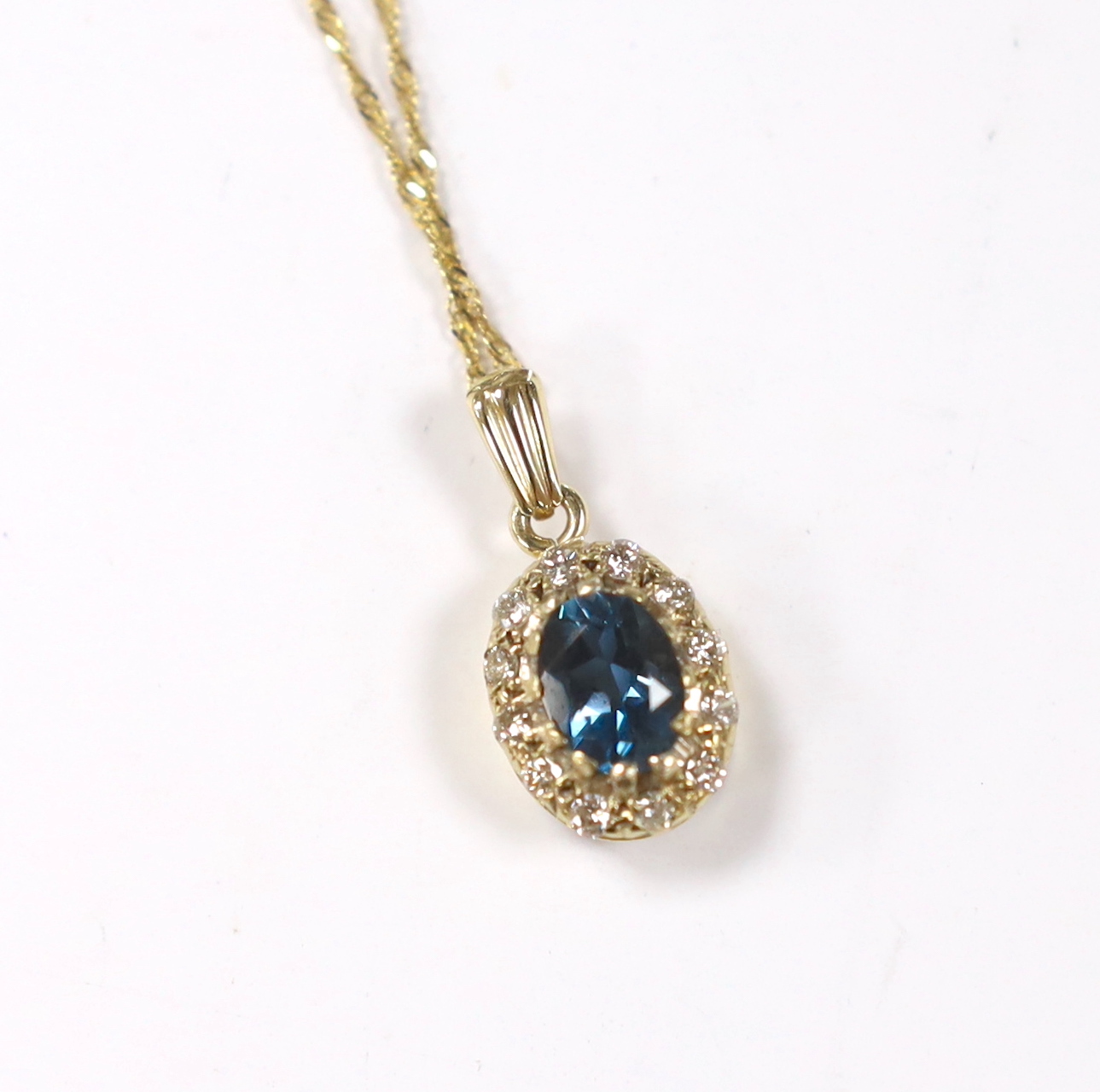 A yellow metal, blue topaz and diamond set oval cluster pendant, 11mm, on a 375 fine link chain, 38cm, gross weight 3 grams.